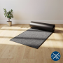 Tapis de passage gris et noir roulé à terre dans une pièce lumineuse, plancher en bois et plante verte en arrière-plan.
