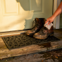 Paillasson extérieur en caoutchouc noir, motif cœur, posé sur une marche avec bottes boueuses; main nettoyant le paillasson.