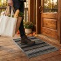 Tapis d'entrée noir et blanc à motifs abstraits, avec rebords gris, posé sur une terrasse en bois devant une porte en bois; une