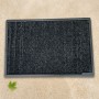 Tapis extérieur astroturf noir 60x90 cm posé sur un sol clair, bordures grises et logo Astroturf en coin. Une feuille verte au c