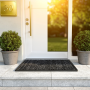 Tapis extérieur astroturf noir 60x90 cm posé devant une porte d’entrée blanche, entouré de pots ronds et d’un paillasson noir.