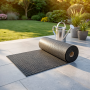 Tapis extérieur sécurisé et drainant en caoutchouc octogonal déroulé sur une terrasse, jardin en arrière-plan et arrosoir en mét