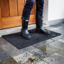 Tapis extérieur noir à mailles octogonales en caoutchouc, posé devant une porte; bottes imperméables sur la surface drainante et
