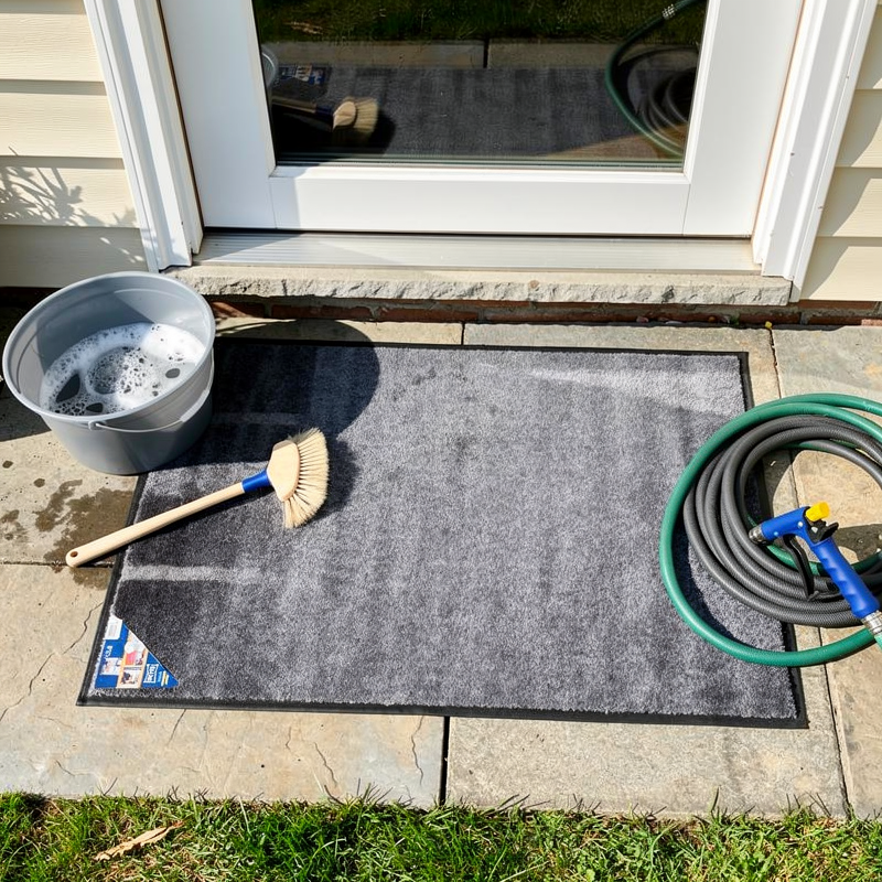 Tapis d'entrée gris sur une terrasse avec seau et brosse pour nettoyage manuel